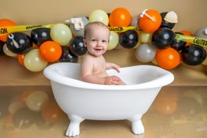 Smiling baby sitting in a white clawfoot bathtub surrounded by orange, black, and gold balloons with caution tape backdrop.