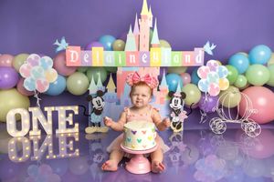 Baby girl sits by a Disney castle cake smash setup with Mickey Mouse, balloons, and Delaineland sign for her first birthday.