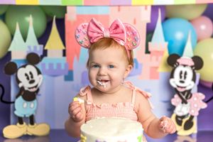 Baby girl in pink bow ears smashes first birthday cake at Disney-themed photo shoot with Mickey and Minnie Mouse.