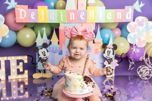 Baby girl smashes first birthday cake at Disneyland-themed party with colorful balloons and Mickey Mouse decorations.