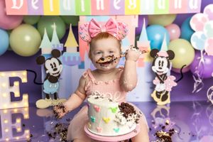 Baby girl in pink tutu smashes first birthday cake at Disney-themed party with Mickey and Minnie Mouse decorations.