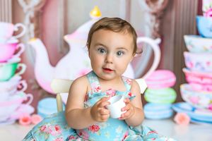 Baby girl in floral dress at tea party themed photo shoot with colorful macarons and teacups backdrop.