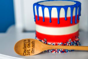 Red, white, and blue drip cake with sprinkles beside a wooden spoon engraved with Caden's 1st Cake Smash.