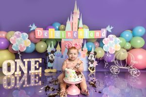 Baby girl celebrates first birthday with Disney-themed cake smash, featuring castle backdrop, balloons, and Mickey Mouse decorations.