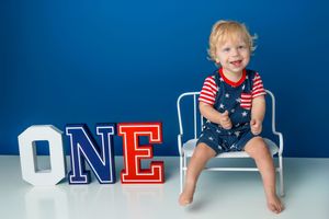Smiling toddler boy sitting on white chair beside colorful ONE letters, wearing patriotic outfit, blue background.