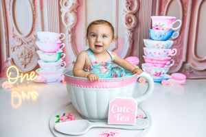 Baby girl sits in giant teacup prop with stacked floral cups, wearing floral romper at One birthday tea party photoshoot.