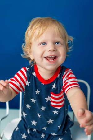 Smiling toddler with blonde curly hair and blue eyes wearing a patriotic stars and stripes outfit against a blue background.