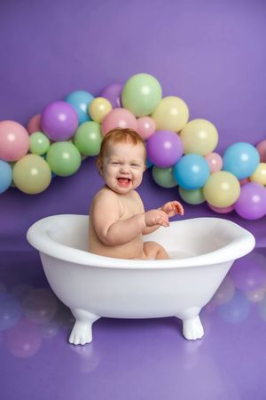 Happy baby sitting in a white mini bathtub laughing, surrounded by colorful pastel balloons on a purple background.