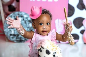 Baby girl in pink outfit and cowboy hat smashes first birthday cake, hands raised with frosting.