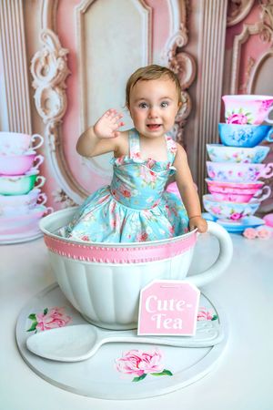 Baby girl in floral dress sitting in giant teacup prop with stacked colorful cups, waving at camera.