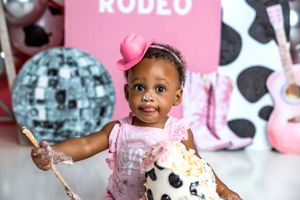 Baby girl in pink outfit and cowboy hat smashes first birthday cake at rodeo-themed photo shoot.