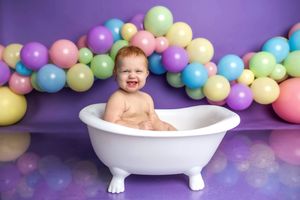 Laughing baby sitting in a white mini bathtub surrounded by colorful pastel balloons on a purple background.