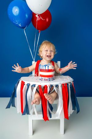 Laughing baby in patriotic red, white, and blue outfit celebrates first birthday with cake and balloons.
