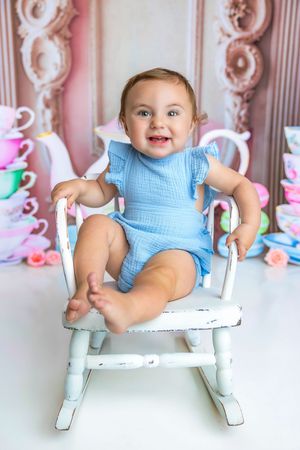 Happy baby in blue romper sitting in white rocking chair with colorful tea cups backdrop.