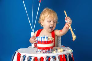 Toddler with curly blond hair joyfully smashing a red, white, and blue first birthday cake with a wooden spoon.