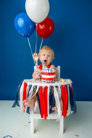 Surprised toddler at first birthday cake smash with red, white, and blue balloons and decorated high chair.