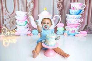 Baby in teapot hat sits by smash cake surrounded by pastel tea cups and macarons at a whimsical first birthday.