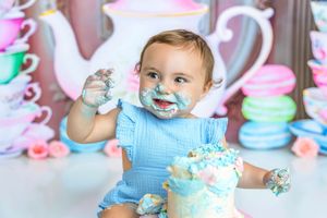Adorable baby enjoying first birthday cake smash in blue outfit with frosting on face and hands.