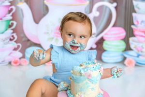 Baby girl in blue romper smashing first birthday cake with frosting on her face, tea party backdrop.