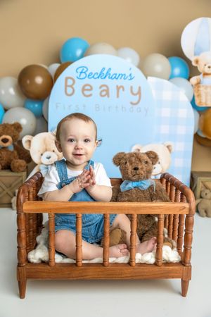 Baby boy claps hands in wooden crib with teddy bear at Beckham's Beary First Birthday photo session.