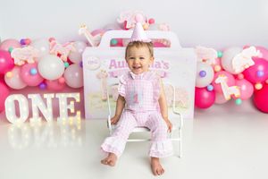 Happy toddler in pink pajamas celebrating first birthday with balloons and ONE sign.