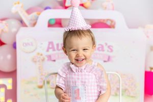Smiling baby girl in pink gingham dress and party hat celebrates first birthday with decorations and balloons behind her.