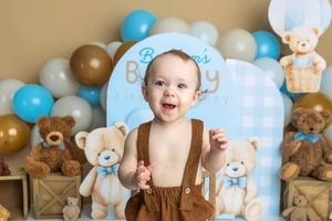A smiling baby in brown suspenders poses with teddy bears and blue and gold balloons at a first birthday cake smash.
