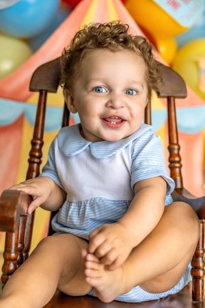 Happy baby with curly hair and blue eyes sits in wooden chair, wearing blue striped outfit at colorful birthday party.
