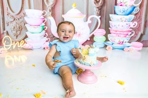 Baby girl in blue dress smiles at her first birthday cake smash with pastel tea party decorations.