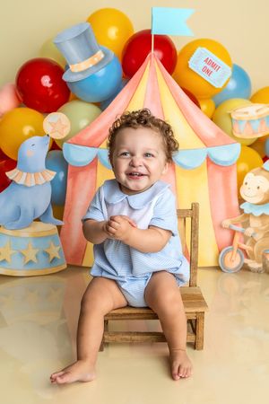 Smiling baby in blue romper sits on wooden chair before colorful circus tent backdrop with balloons for first birthday.