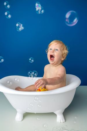 Delighted toddler sitting in white clawfoot bathtub with rubber duck, surrounded by floating soap bubbles on blue background.