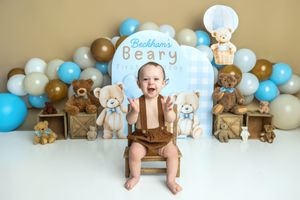 Baby boy sits on a chair surrounded by teddy bears and balloons at a Beary first birthday cake smash photo session.