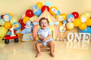 Baby boy smiles at circus-themed first birthday cake smash with colorful balloons and ONE sign backdrop.