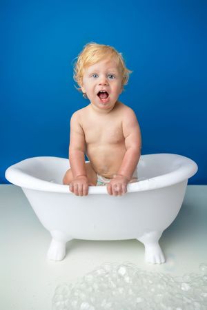 Surprised blonde toddler sitting in a small white clawfoot bathtub with bubbles against a blue background.