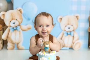 Smiling baby boy at first birthday cake smash with teddy bear theme, blue backdrop, and decorated cake.