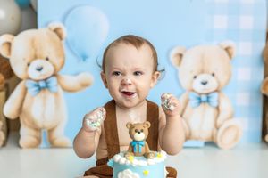 Baby boy smiling at first birthday cake smash with teddy bear decorations and blue balloon backdrop.