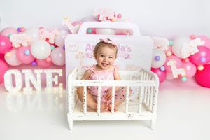 Smiling baby girl in pink stands in white crib at Aubriella's first birthday ONE balloon backdrop session.
