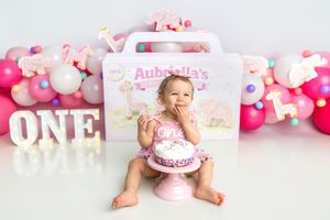 Baby girl in pink romper sits before smash cake at pink ONE birthday setup with balloon garland.