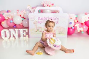 Baby girl in pink romper holds smashed birthday cake at Aubriella's ONE themed first birthday shoot.