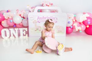 Baby girl in pink romper examines smashed birthday cake at colorful ONE first birthday photo session.