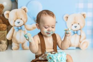 Baby boy in brown suspenders smashes first birthday cake, surrounded by teddy bear decorations on blue backdrop.