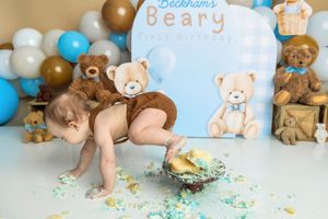 Baby enjoys a teddy bear themed first birthday cake smash with blue balloons and stuffed bears in the background.