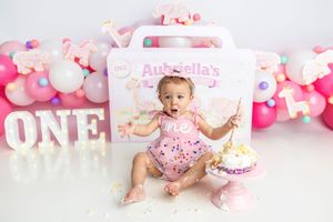 Surprised baby girl holds cake topper at pink first birthday smash cake session with balloon decor.