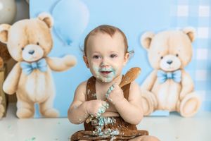 Baby boy enjoys first birthday cake smash with teddy bear backdrop, covered in blue frosting and chocolate cake.