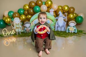 Baby in Hobbit costume celebrates first birthday with Lord of the Rings themed backdrop, balloons, and One Ring prop.