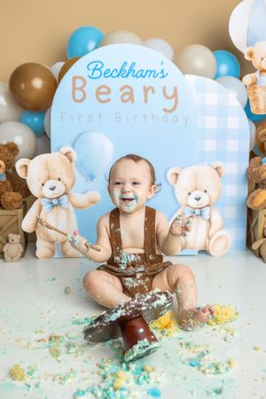 Baby boy smiles during Beckham's Beary First Birthday cake smash with teddy bear backdrop and colorful balloons.