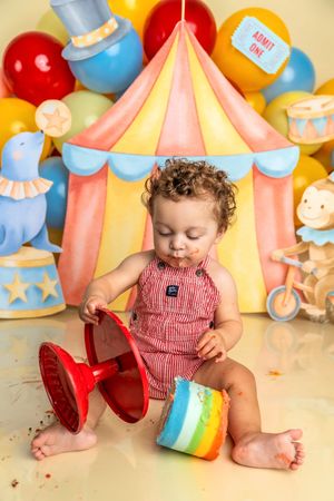 Toddler in red romper enjoys first birthday cake smash with colorful circus tent backdrop and balloons.