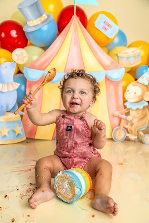 Baby's first birthday cake smash with circus theme, colorful balloons, and tent backdrop, holding a toy mallet.