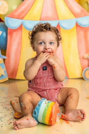 Baby in red romper eating rainbow cake at circus-themed first birthday smash session.