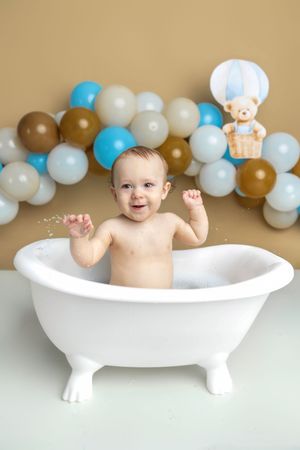 Happy baby splashes in white mini bathtub surrounded by blue, gold, and white balloon garland backdrop.